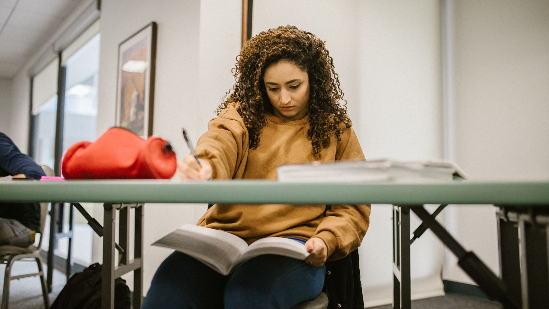 Young woman with curly hair reading a textbook and taking notes at a classroom desk