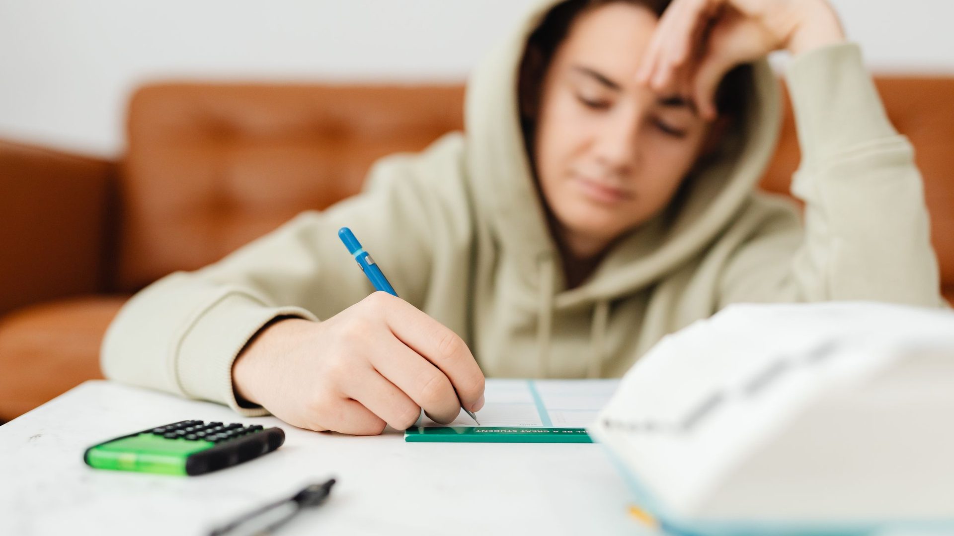 Young person in a hooded sweatshirt writing on a worksheet at a table, with a calculator nearby