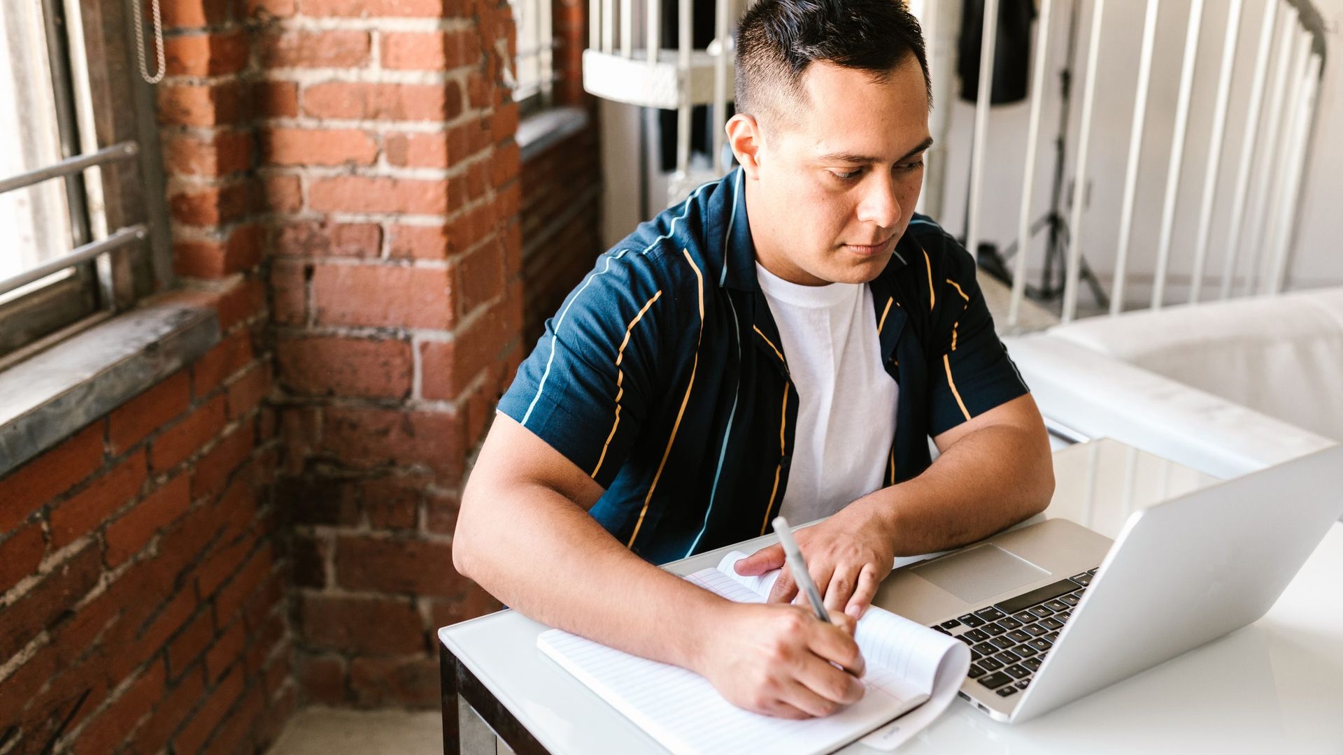 Young man writing in a notebook next to a laptop while studying at a desk by a brick wall