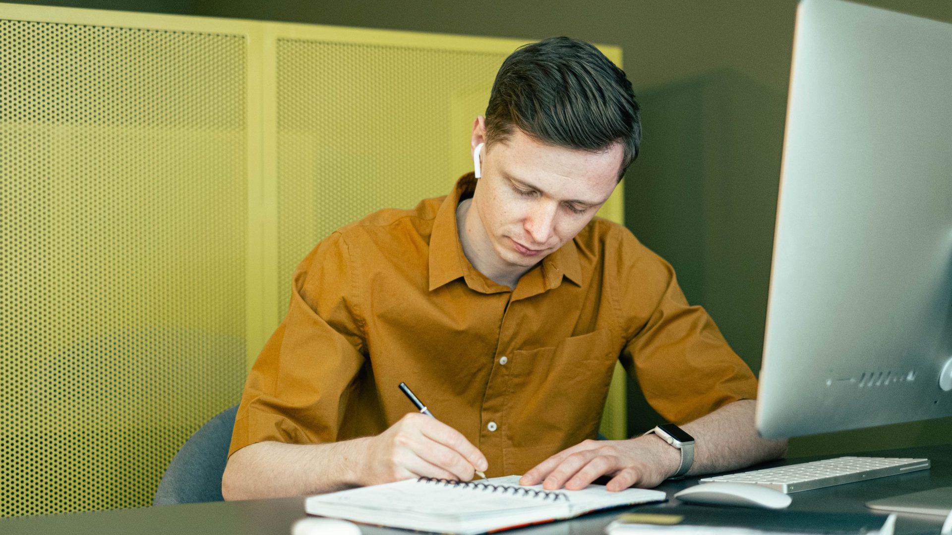 Young man in a mustard shirt writing in a notebook at a desk next to a desktop computer