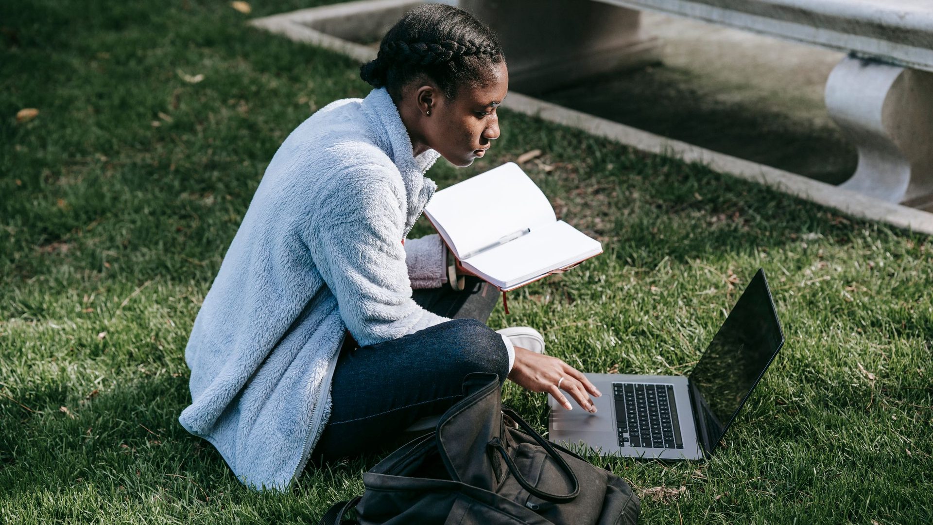 Young woman sitting on a campus lawn with a laptop and an open notebook