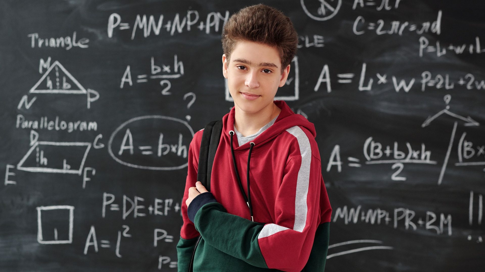 Teenage boy with a backpack standing in front of a chalkboard covered with area formulas