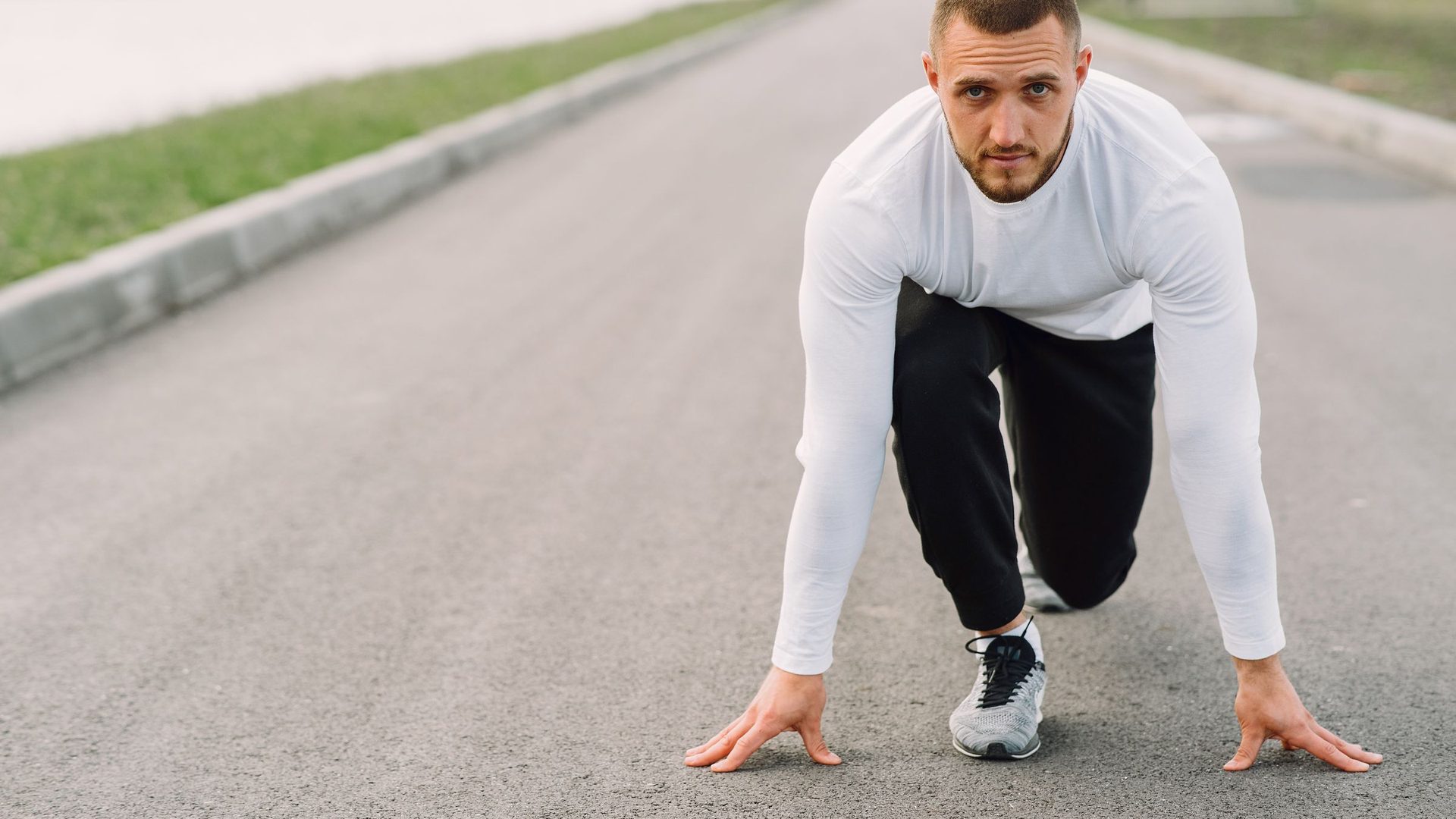 Young man in a white long-sleeve shirt crouched in a sprint start position on an empty road