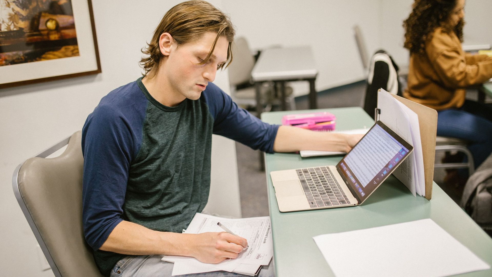 Young man writing on a worksheet beside an open laptop at a classroom desk