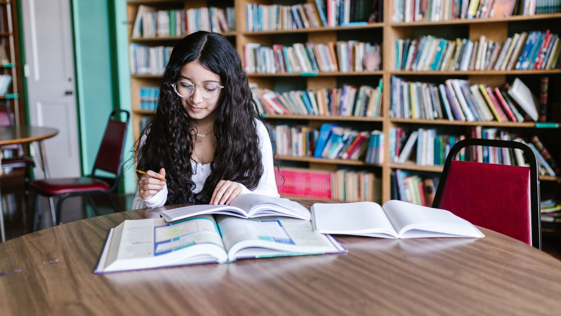 Young woman with long curly hair and glasses studying open textbooks at a library table