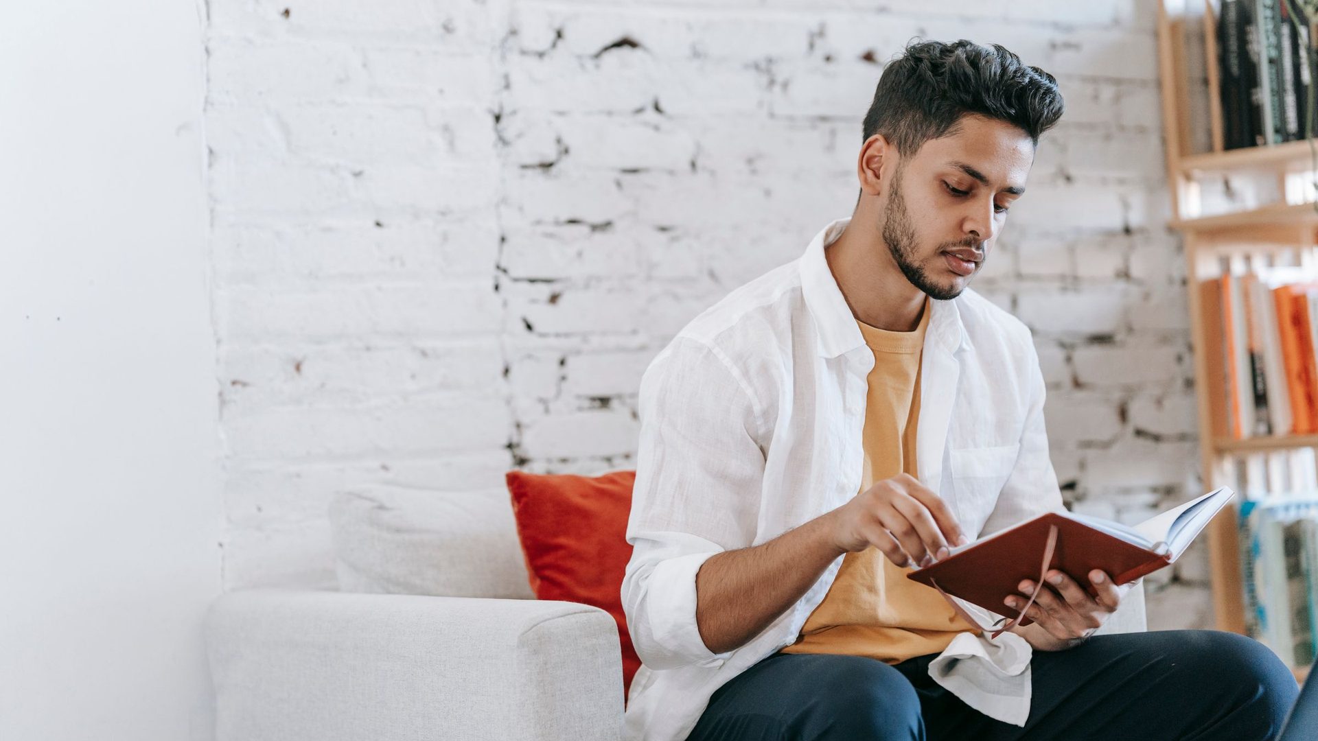Young man in a white shirt reading a small notebook on a couch against a white brick wall