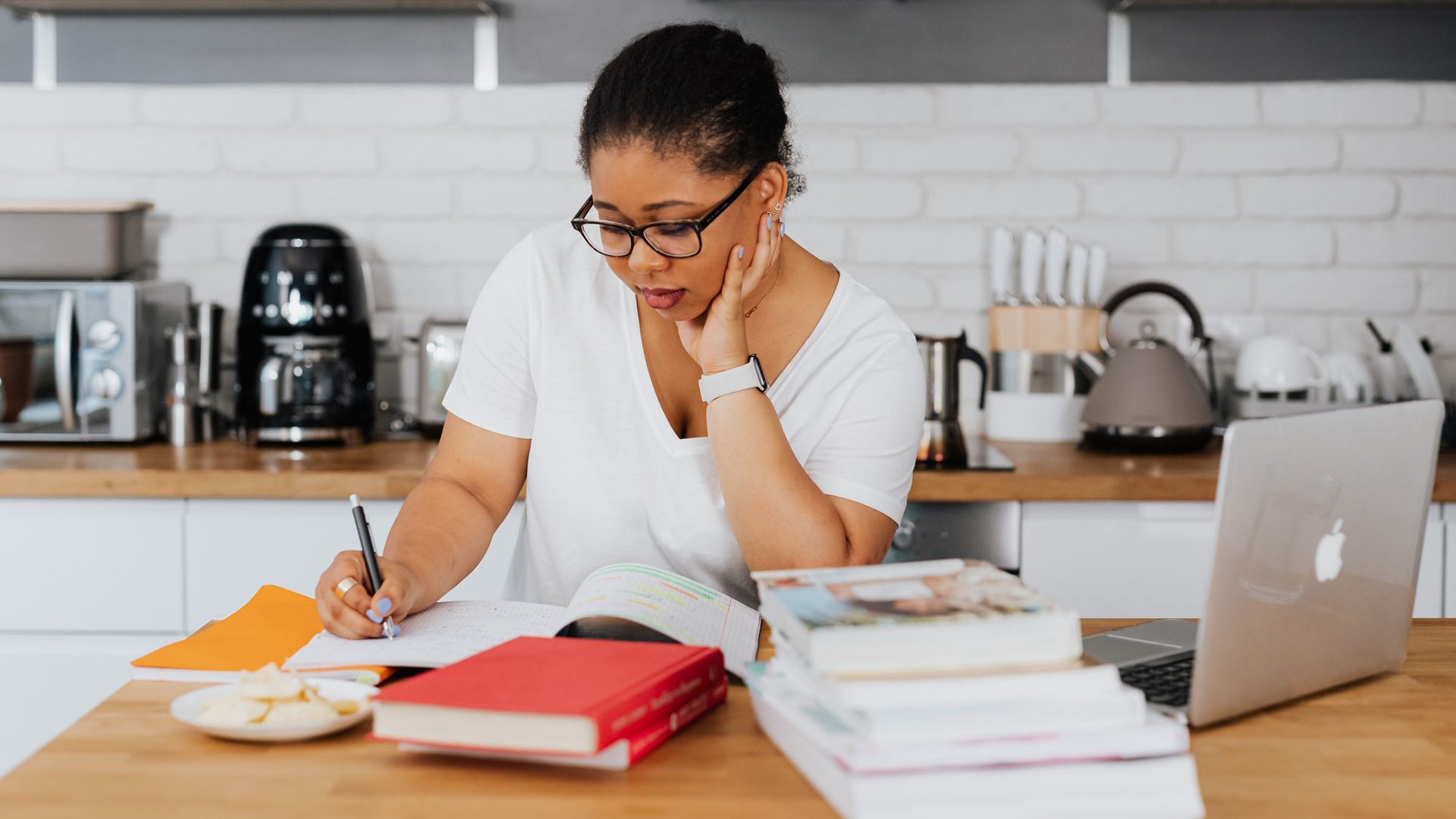 Woman in glasses writing in a notebook at a kitchen counter with a stack of books and a laptop