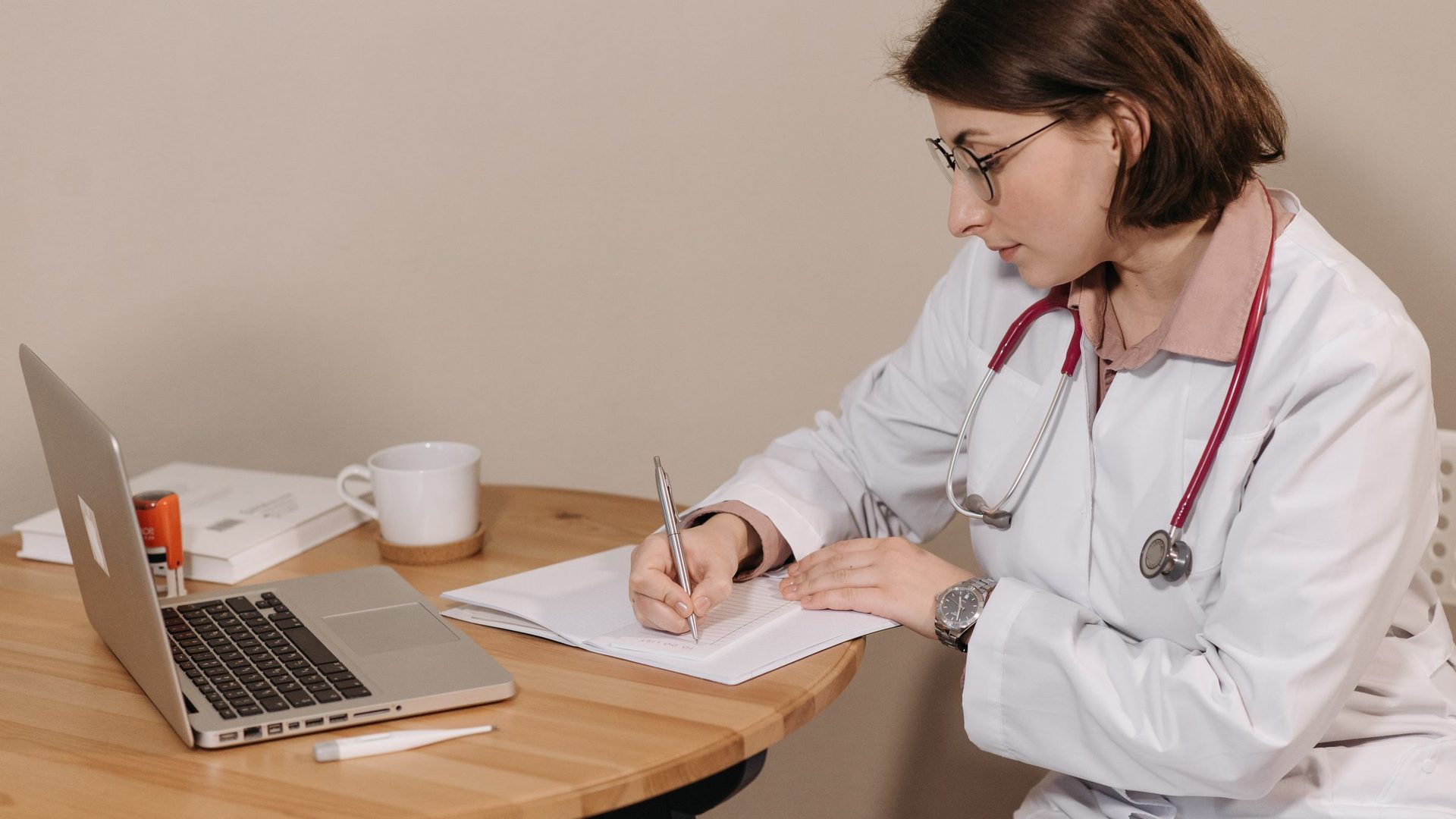 Woman in a white coat with a stethoscope writing notes at a small table next to a laptop