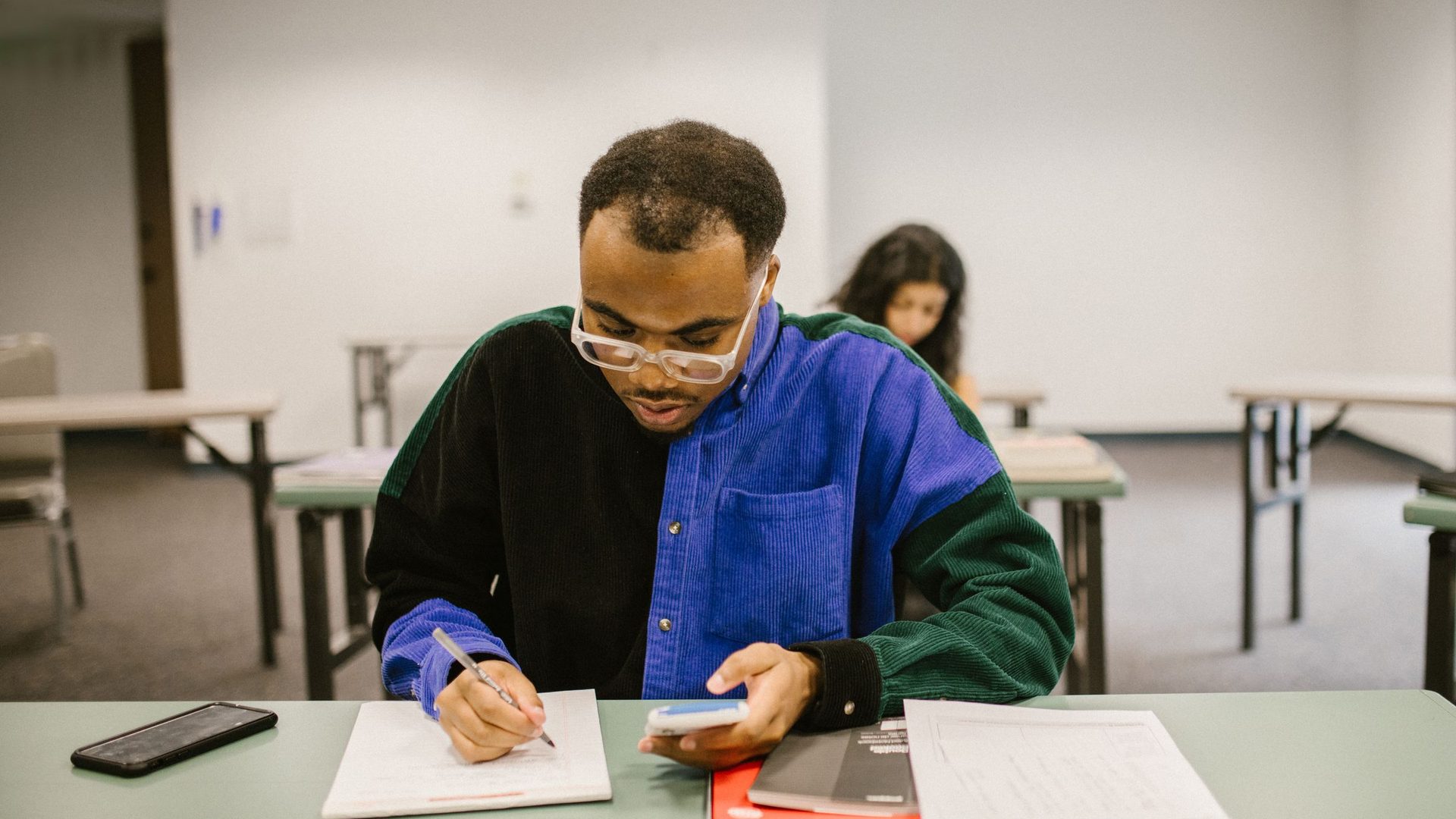 Man in glasses and a color-block jacket writing on a worksheet at a desk in an adult education classroom