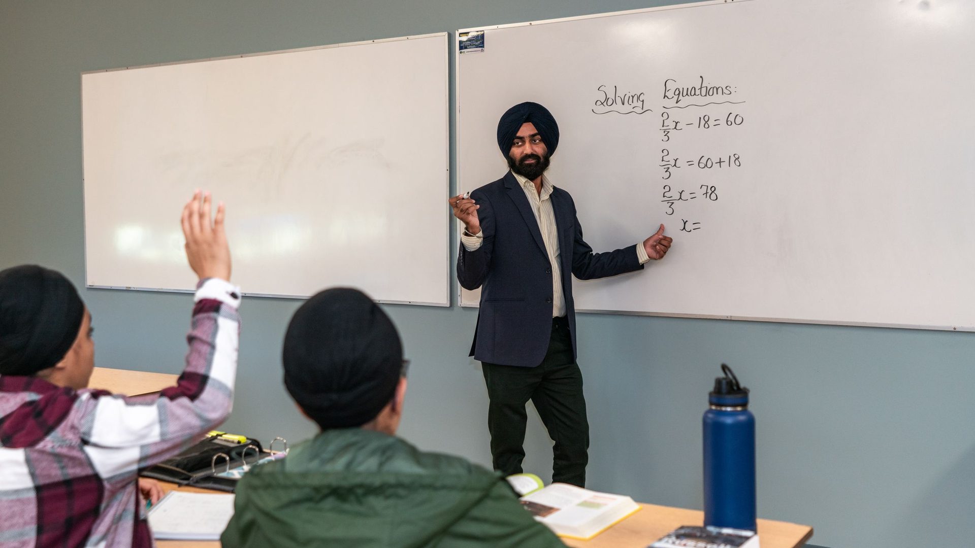 Teacher pointing at equations on a whiteboard while a student in the front row raises a hand