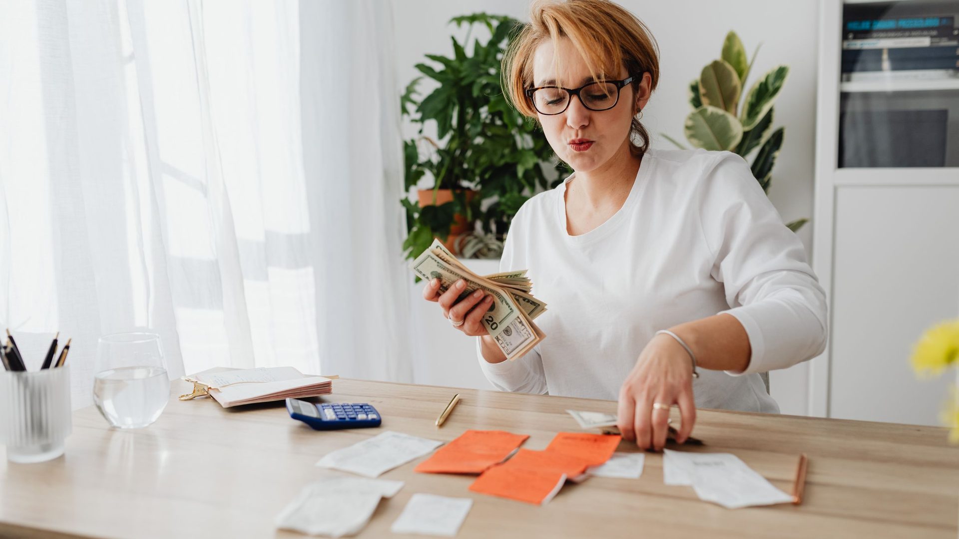 Woman in glasses counting cash at a table covered with receipts and a calculator