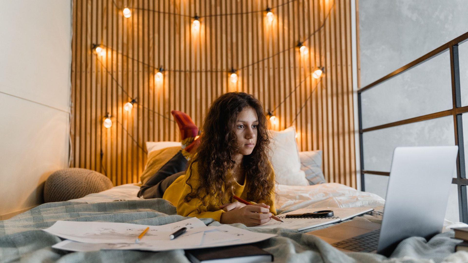 Teenage girl with curly hair studying on her bed with notes and a laptop under string lights