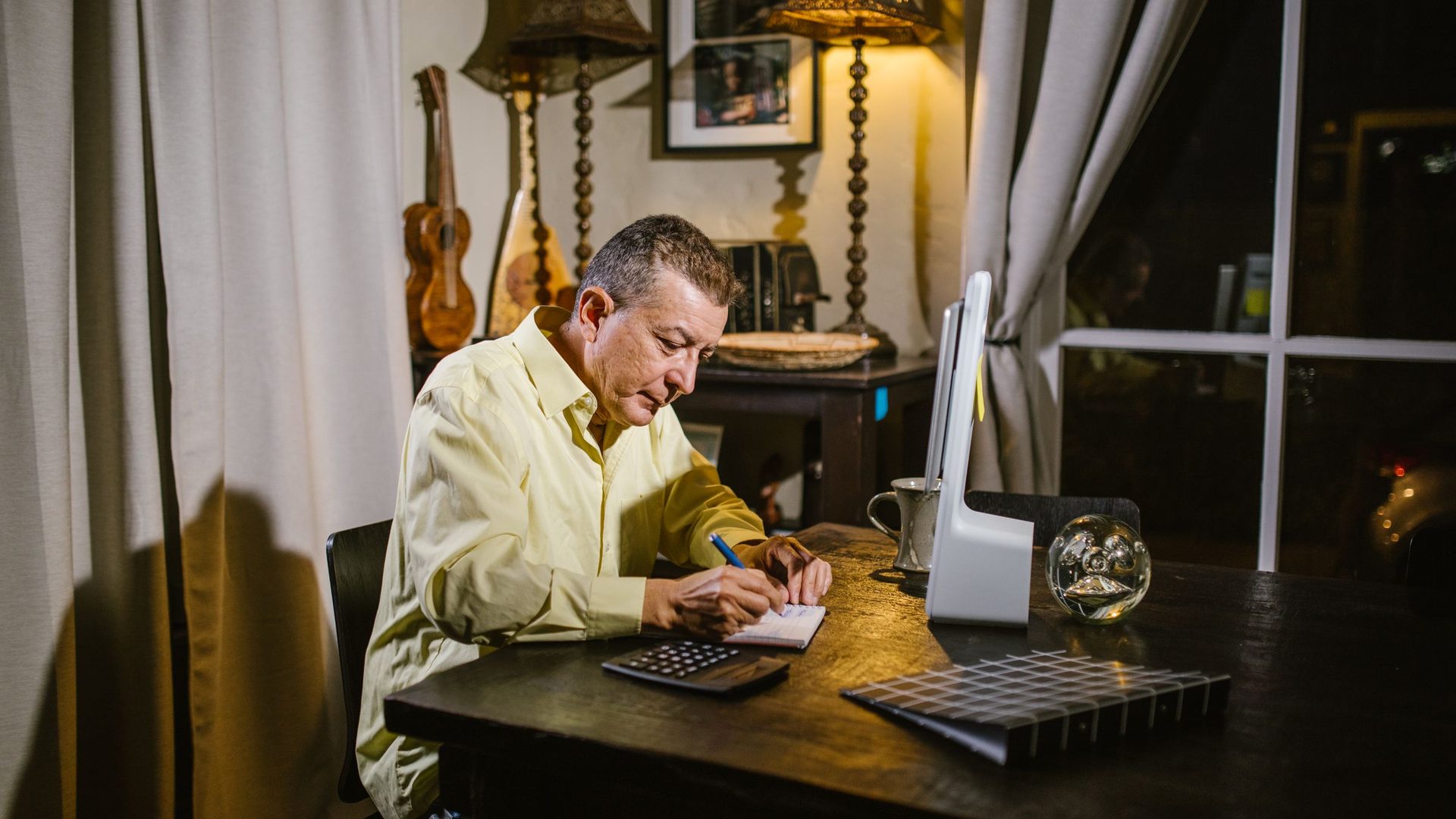 Older man in a yellow shirt writing notes at a wooden desk at home in the evening