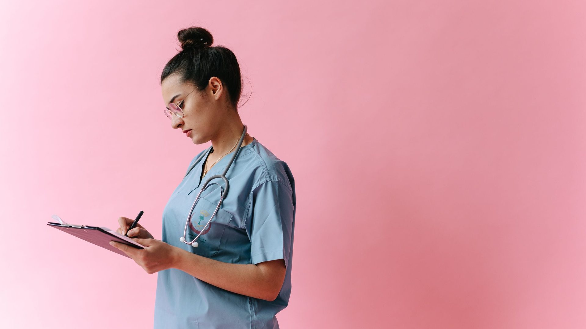 Young woman in blue scrubs with a stethoscope writing on a clipboard against a pink background