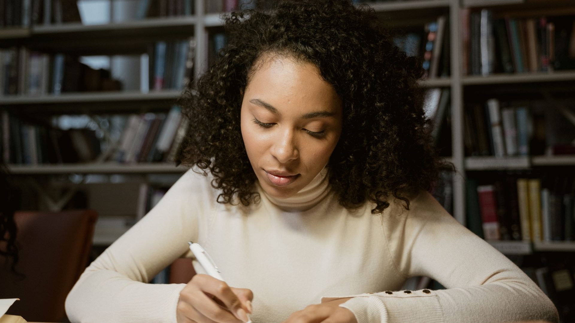 Young woman with curly hair writing notes at a desk in front of library bookshelves