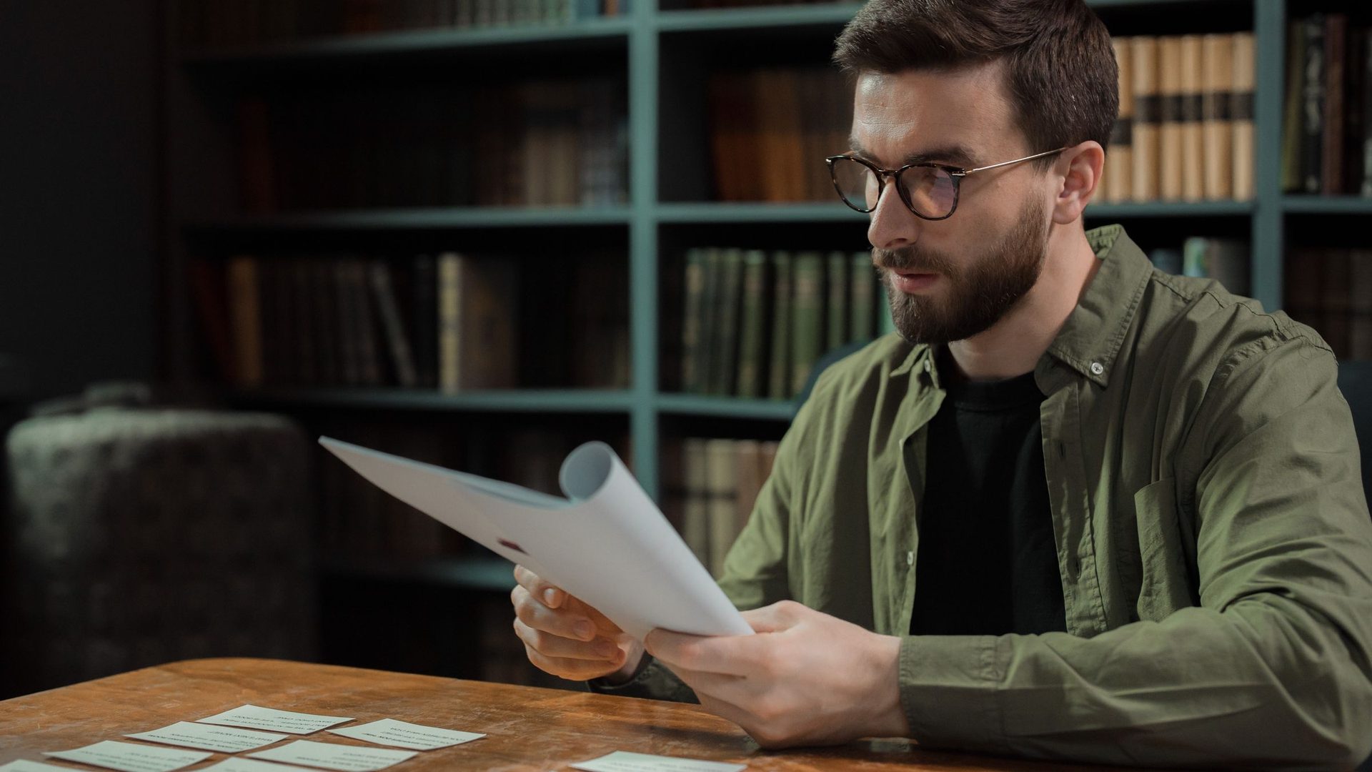 Bearded man in glasses reading a sheet of paper at a table in a dim library